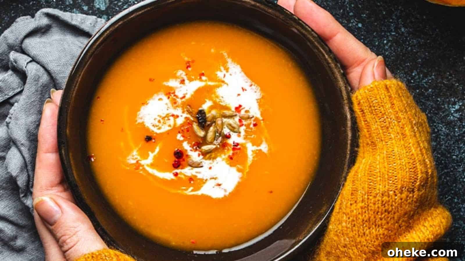 17 Comforting Recipes to Keep You Warm This Season 16 A bowl of coconut pumpkin soup garnished with cream, seeds, and spices is held by two hands in yellow sweaters.