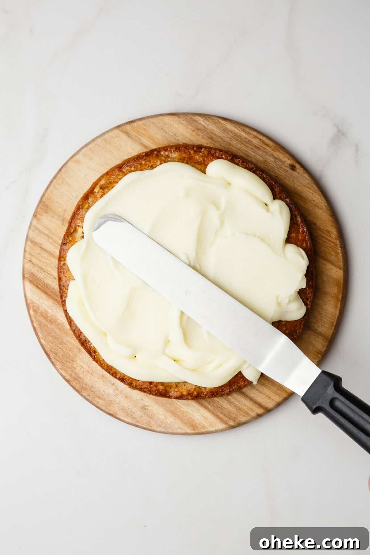 Top Cake Decorating Tools: 12 Must-Have Supplies for Bakers 4 top down image of an off set spatula frosting a vanilla cake sitting on a round wooden board.