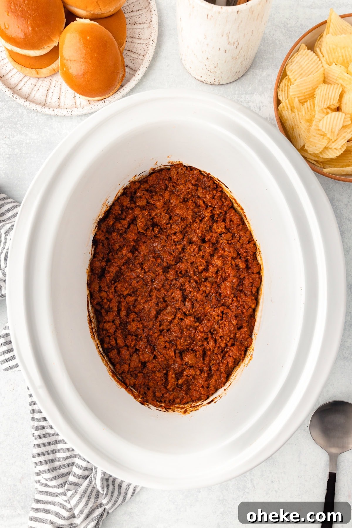 Slow Cooker Sloppy Joes Recipe for Tender, Flavorful Sandwiches 5 A slow cooker filled with cooked sloppy joe meat sits on a white surface, surrounded by a plate of burger buns, a bowl of potato chips, a striped towel, and a large serving spoon.