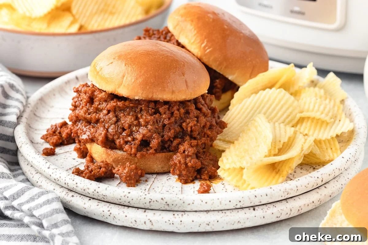 Slow Cooker Sloppy Joes Recipe for Tender, Flavorful Sandwiches 6 Two sloppy joe sandwiches on a plate with a serving of ridged potato chips on the side. Another plate of chips is in the background, along with a striped napkin.