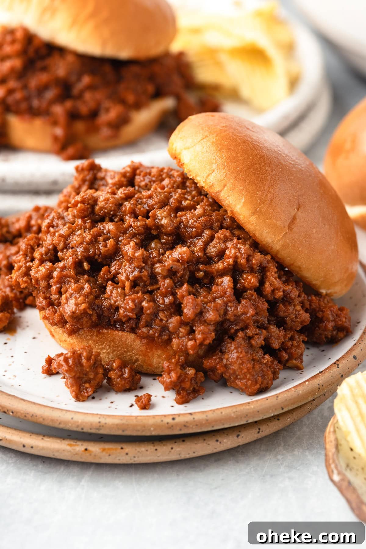 Slow Cooker Sloppy Joes Recipe for Tender, Flavorful Sandwiches 7 A close-up of a sloppy joe sandwich with seasoned ground beef spilling out of a soft bun, served on a plate with potato chips in the background.