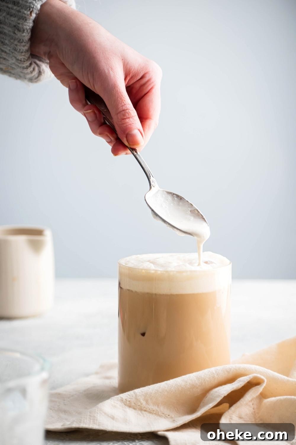 Starbucks Iced White Chocolate Mocha Copycat Recipe — How to Make at Home 2 Hand holding a spoon pouring foam on top of an iced white chocolate mocha in a glass.