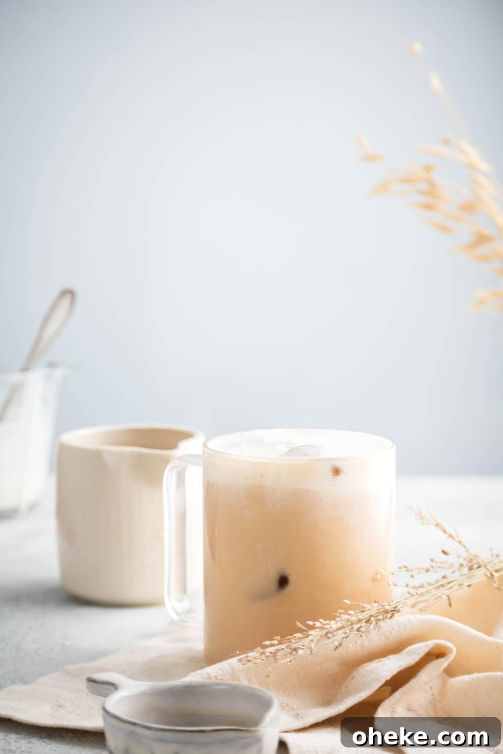 Starbucks Iced White Chocolate Mocha Copycat Recipe — How to Make at Home 3 Part of a small white bowl in front of a white napkin that has a piece of straw on it in front of an iced white chocolate mocha in a glass cup. Behind it is a white pitcher on a gray counter.