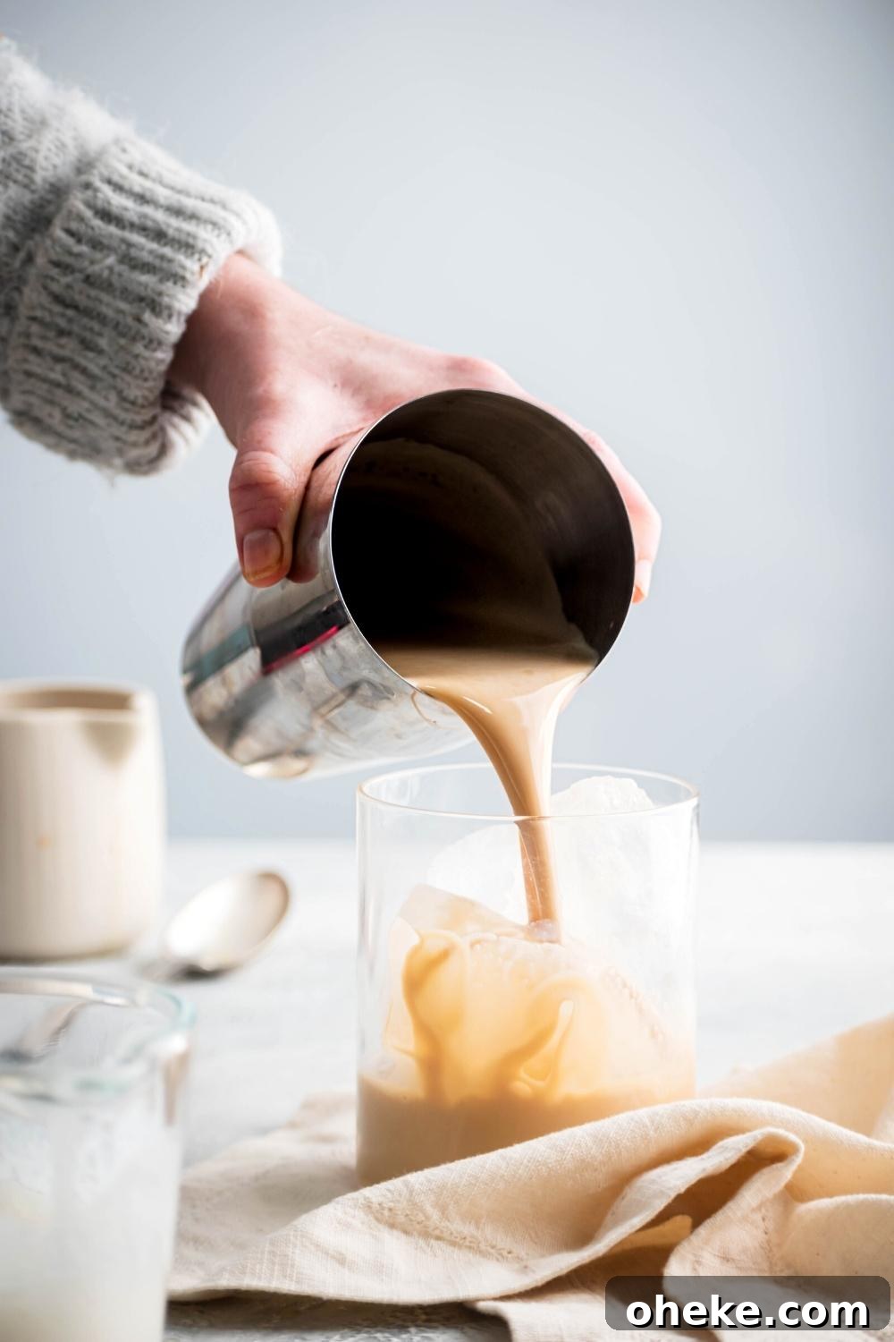 Starbucks Iced White Chocolate Mocha Copycat Recipe — How to Make at Home 6 A hand holding a stainless steel cup pouring an iced white chocolate mocha into a glass top that is filled with ice.