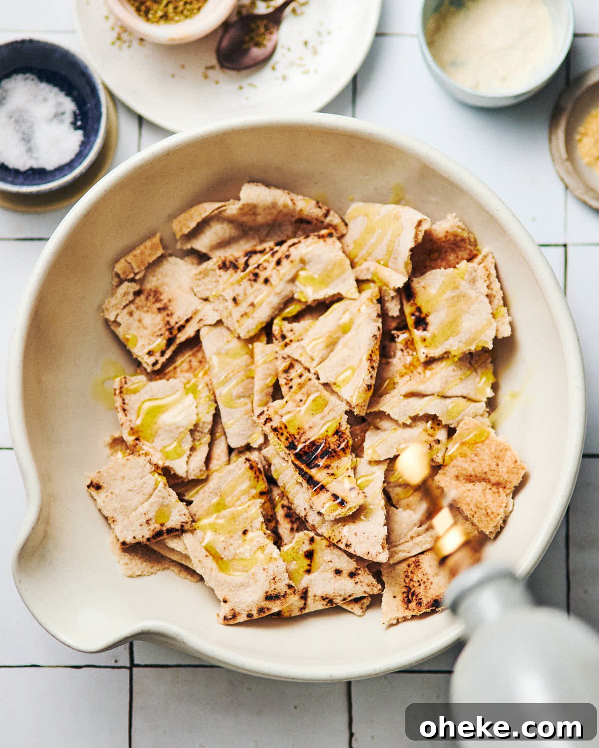 A white ceramic bowl with bite-sized pieces of pita chips, with someone drizzling olive oil on top.