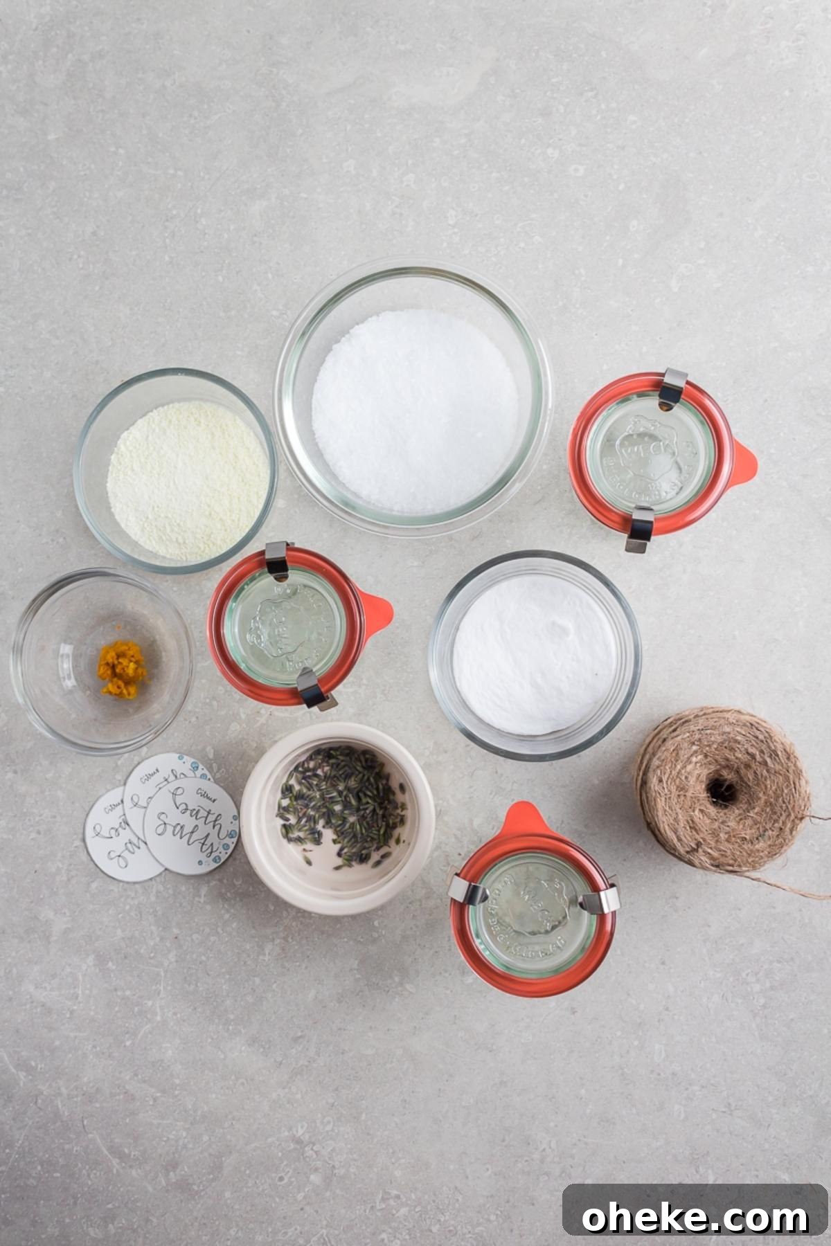 Top view of jars and bowls with DIY Lavender Orange Bath Salts ingredients, twine, tags, and dried lavender on a gray surface.