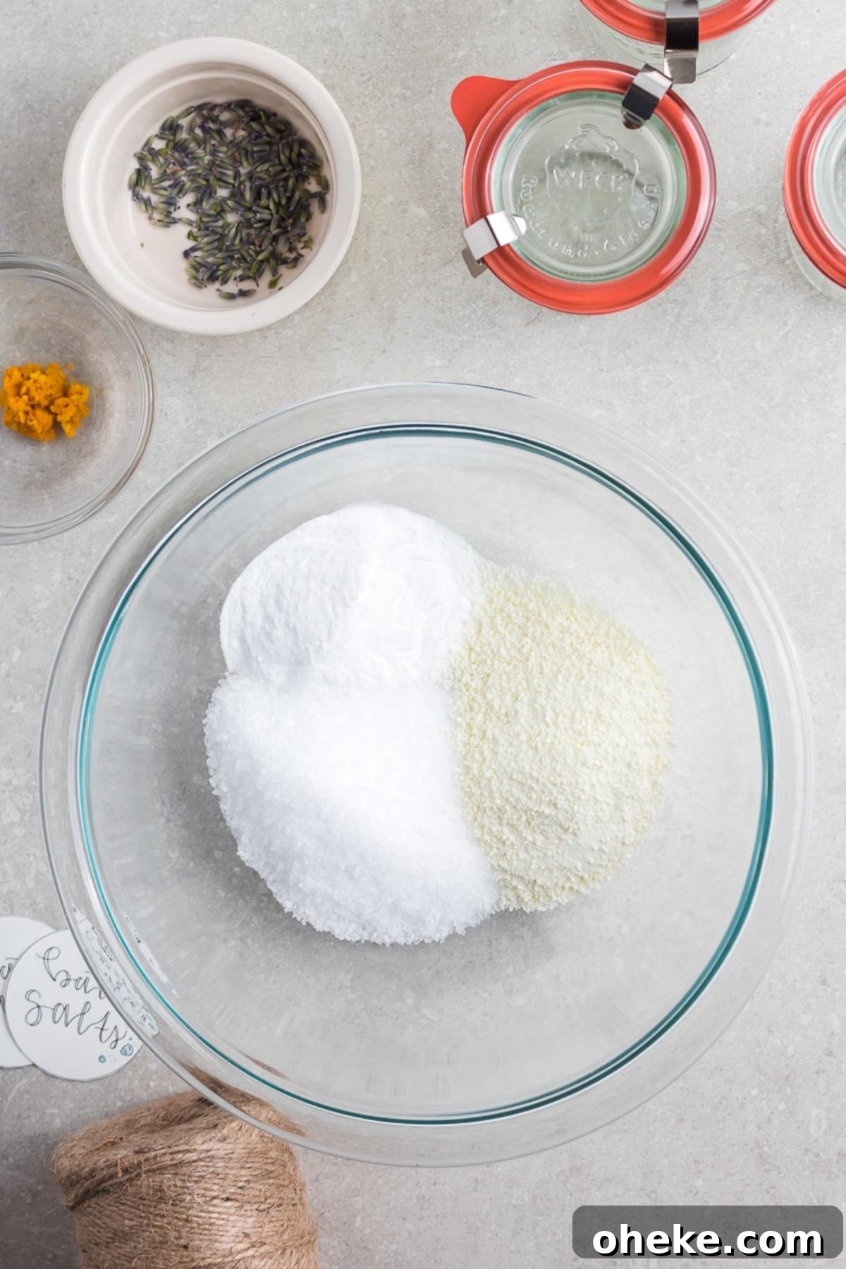 A glass bowl with DIY Lavender Orange Bath Salts—salts and powders beside jars, dried lavender, orange zest, and twine—sits beautifully arranged on a countertop.