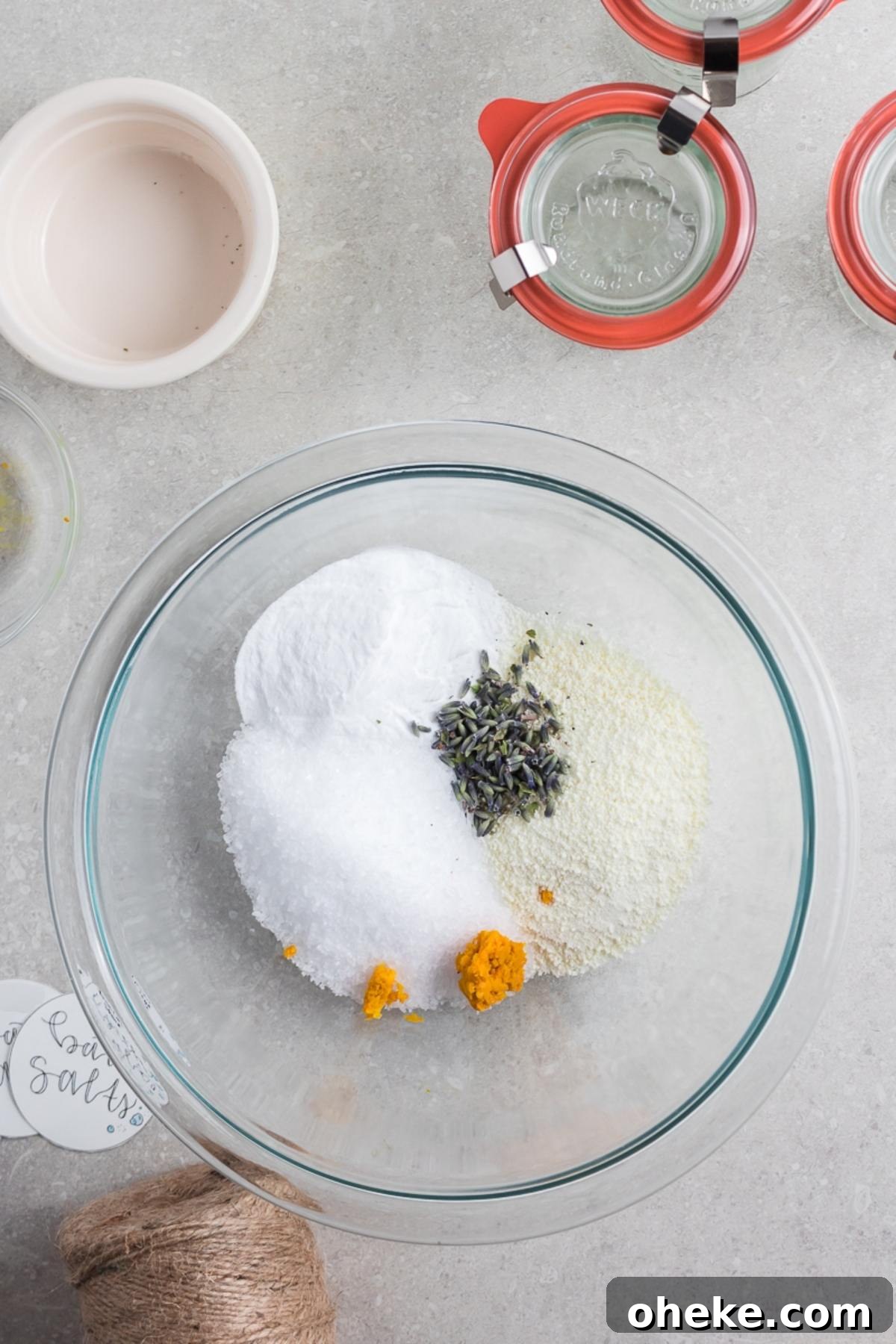 A glass bowl with salt, lavender, and yellow powder sits on a gray counter, surrounded by jars and twine—perfect for making your own DIY Lavender Orange Bath Salts.