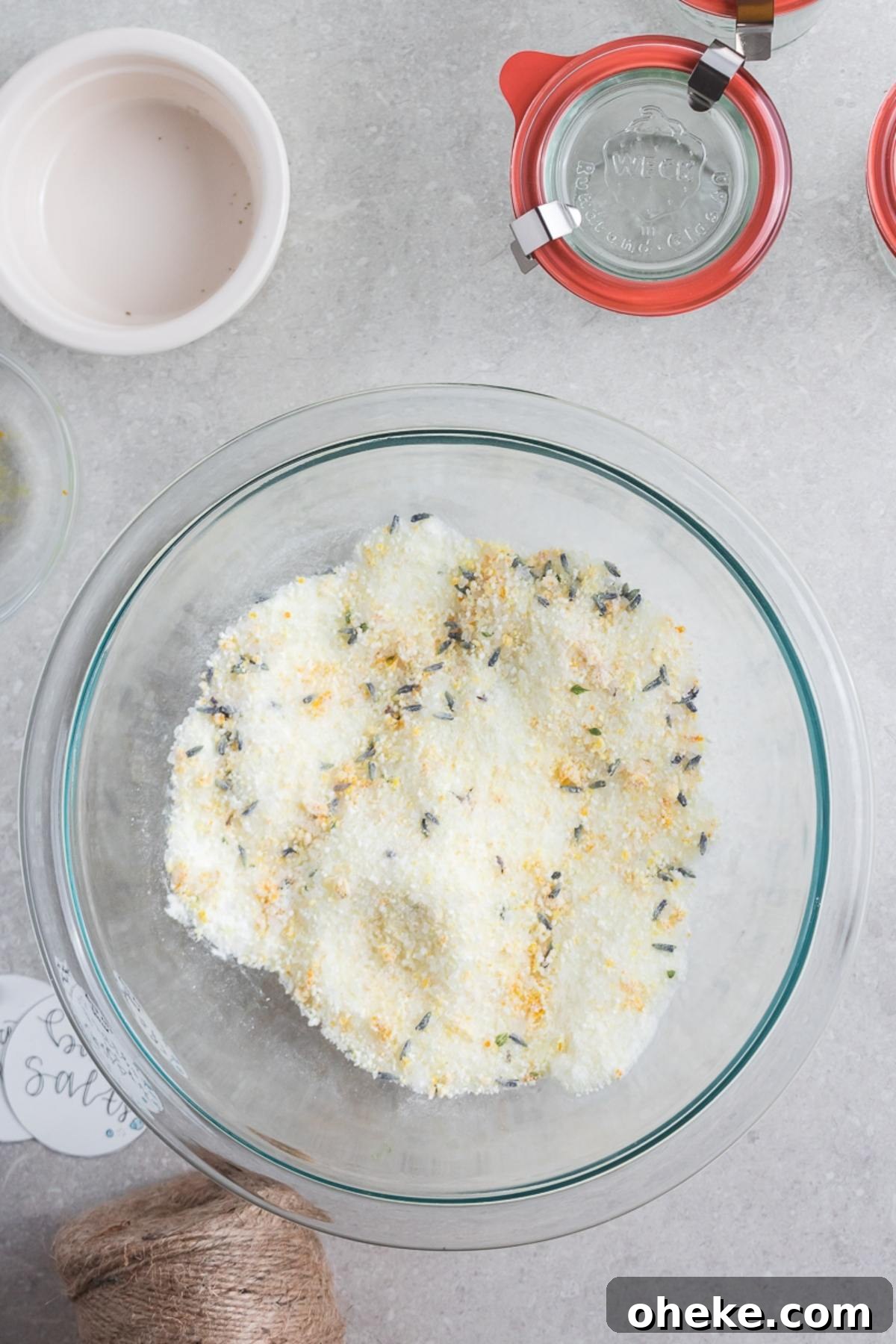 Glass bowl with a mixture of salt, dried lavender, and lemon zest for DIY Lavender Orange Bath Salts on a gray countertop, with jars nearby.