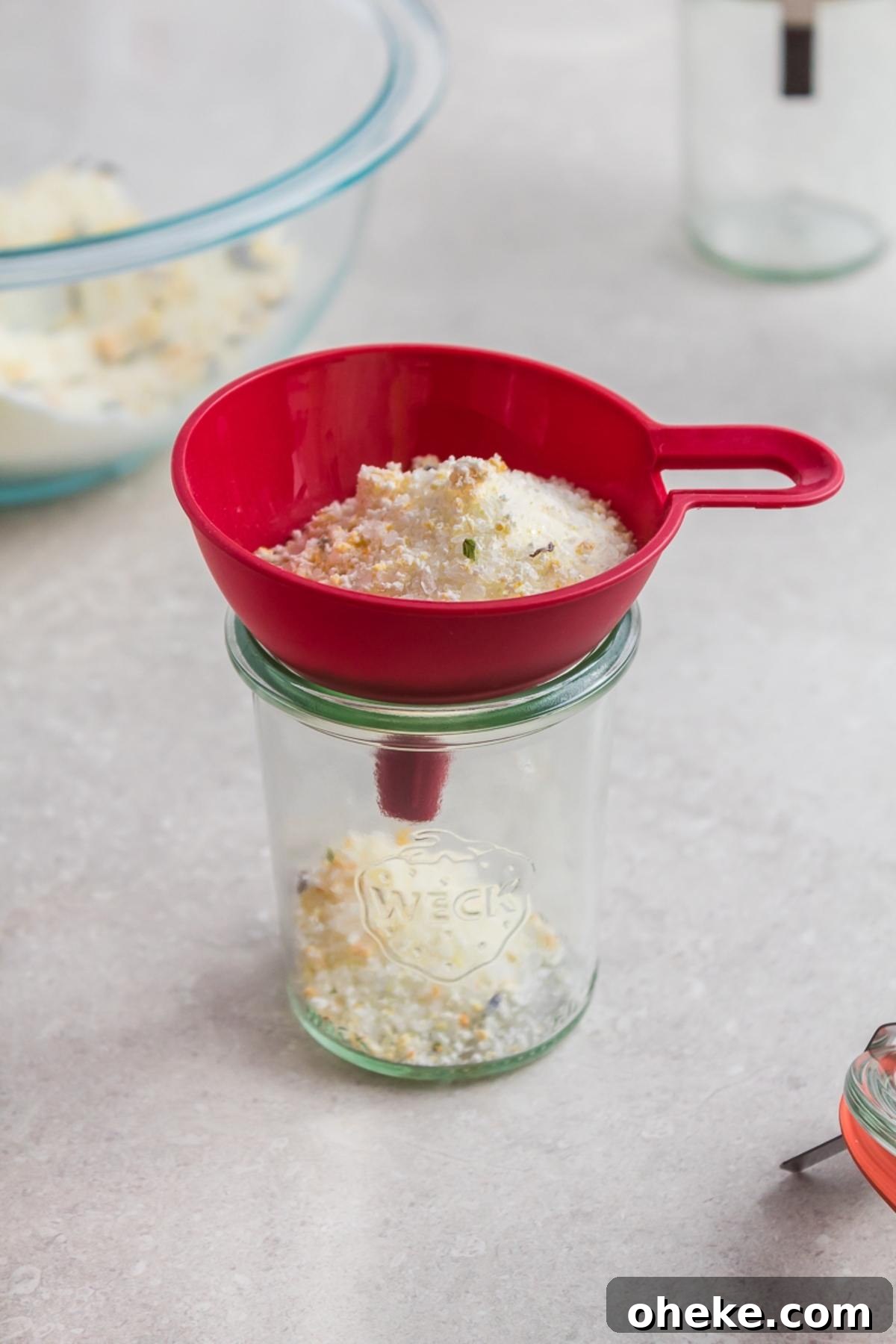 A red funnel pours a crumbly mixture of DIY Lavender Orange Bath Salts into a glass jar on a light countertop.