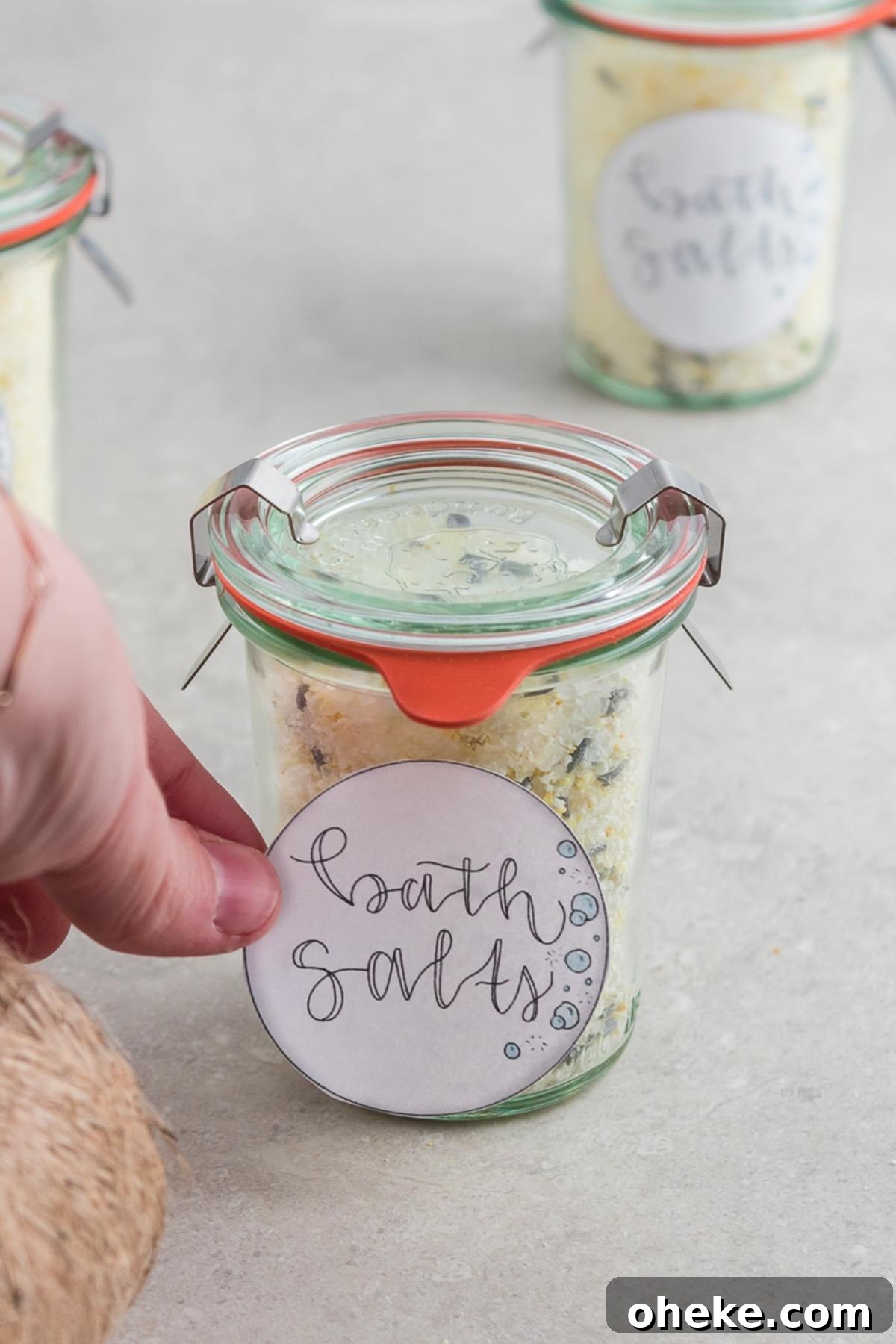 A hand holds a jar labeled "bath salts" filled with homemade DIY Lavender Orange Bath Salts on a light surface.