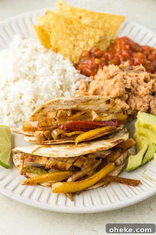 A plate of chicken fajita tacos with grilled bell peppers and onions, served alongside white rice, tortilla chips, salsa, refried beans, sliced avocado, and a lime wedge.