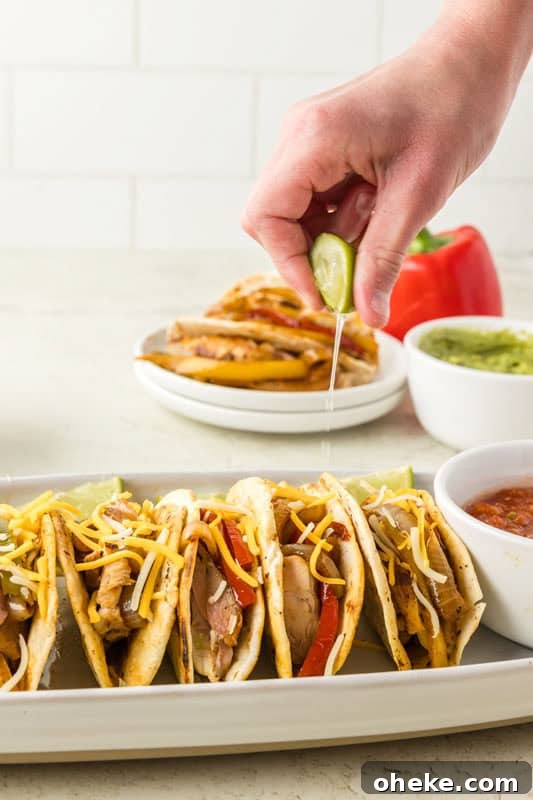 A hand squeezing lime juice over a row of chicken fajita tacos filled with meat, vegetables, and cheese, with side dishes of guacamole and salsa in the background.