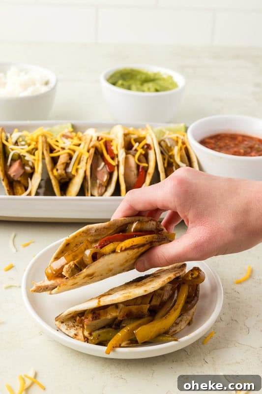 A hand holding a taco filled with grilled vegetables, with a plate of chicken fajita tacos topped with shredded cheese and bowls of rice, guacamole, and salsa in the background.
