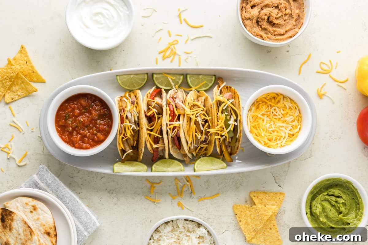 A plate of four chicken fajita tacos with lime wedges. Surrounding the plate are bowls of salsa, shredded cheese, sour cream, beans, guacamole, and tortilla chips. An additional bowl with rice is visible.