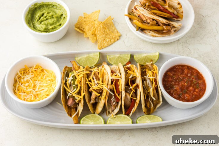 A plate of four chicken fajita tacos topped with shredded cheese and lime slices, accompanied by bowls of shredded cheese, salsa, guacamole, and tortilla chips, with a bowl of grilled vegetables and chicken nearby.