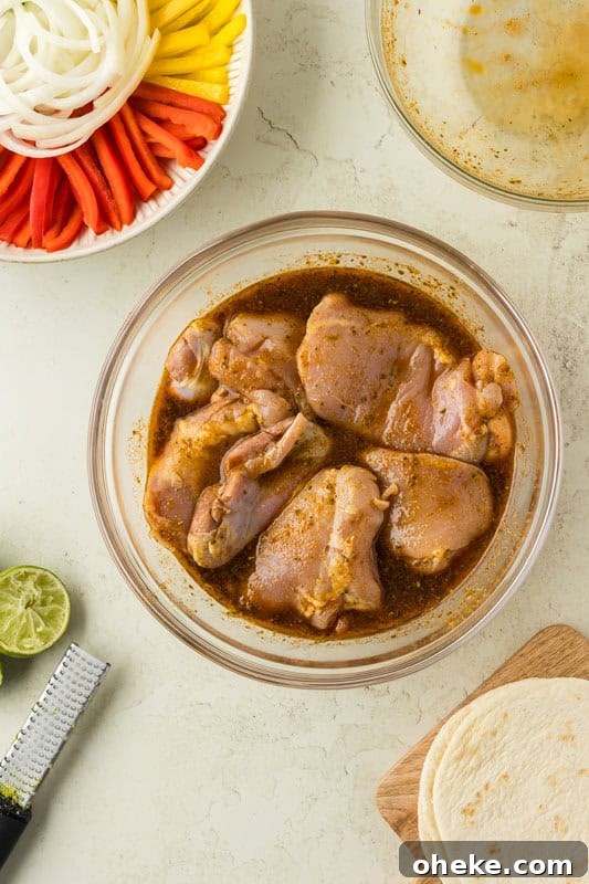 A bowl of raw chicken pieces marinating in a brown sauce sits on the counter, ready to become delicious chicken fajita tacos. Nearby, a dish of sliced vegetables, halved limes, a zester, tortillas on a cutting board, and an empty dish await their turn in this flavorful feast.