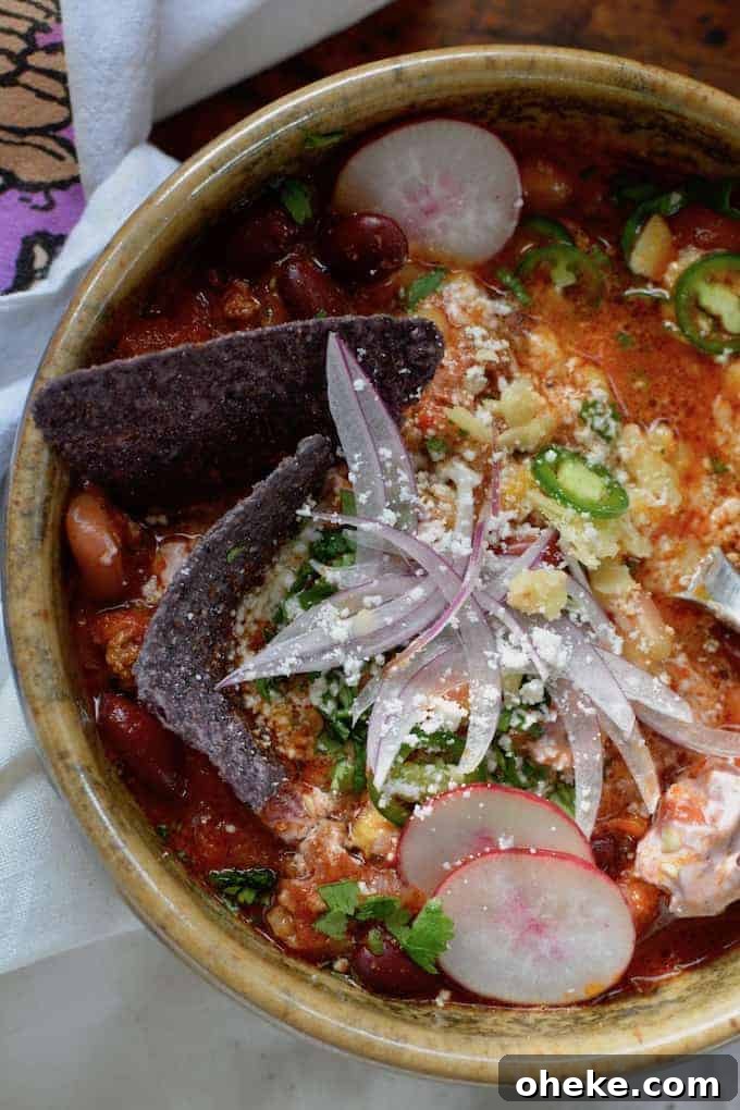 A bowl of chili with beans, radish slices, red onions slices, serrano Chile slices, and a crumbling of white cheese on top.