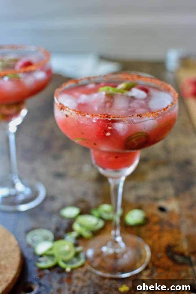 Two pink margaritas sitting on a wood table with slices of jalapeno, fresh raspberries, and ice floating in the glasses.