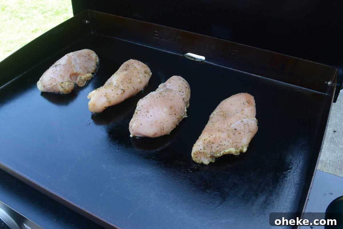 Four uncooked chicken breast pieces with seasoning on a griddle 