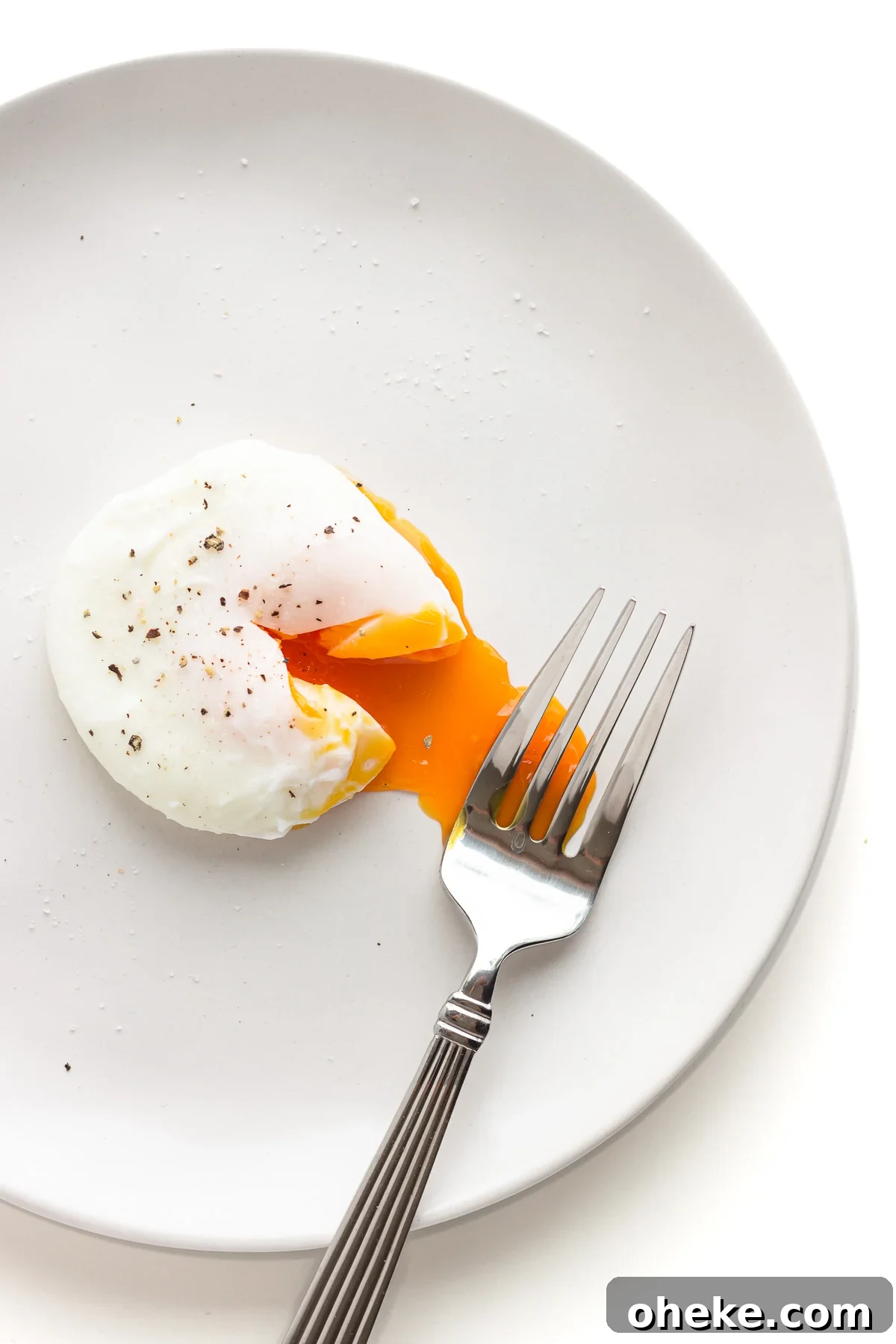 A single poached egg that has been cut into to release the oozy yolk on a white plate with fork.